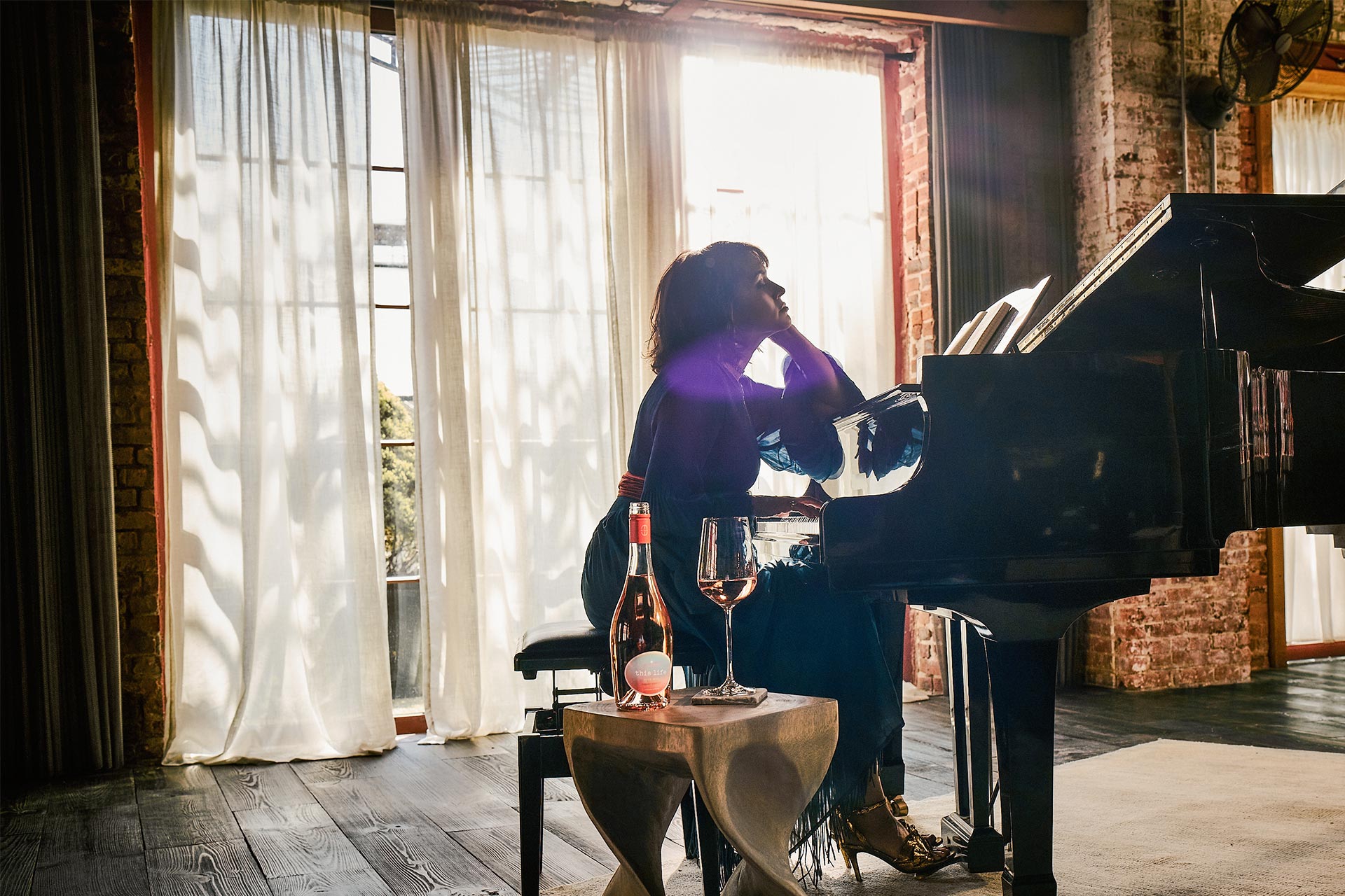 Norah Jones sitting at piano with a bottle and glass of This Life wine.